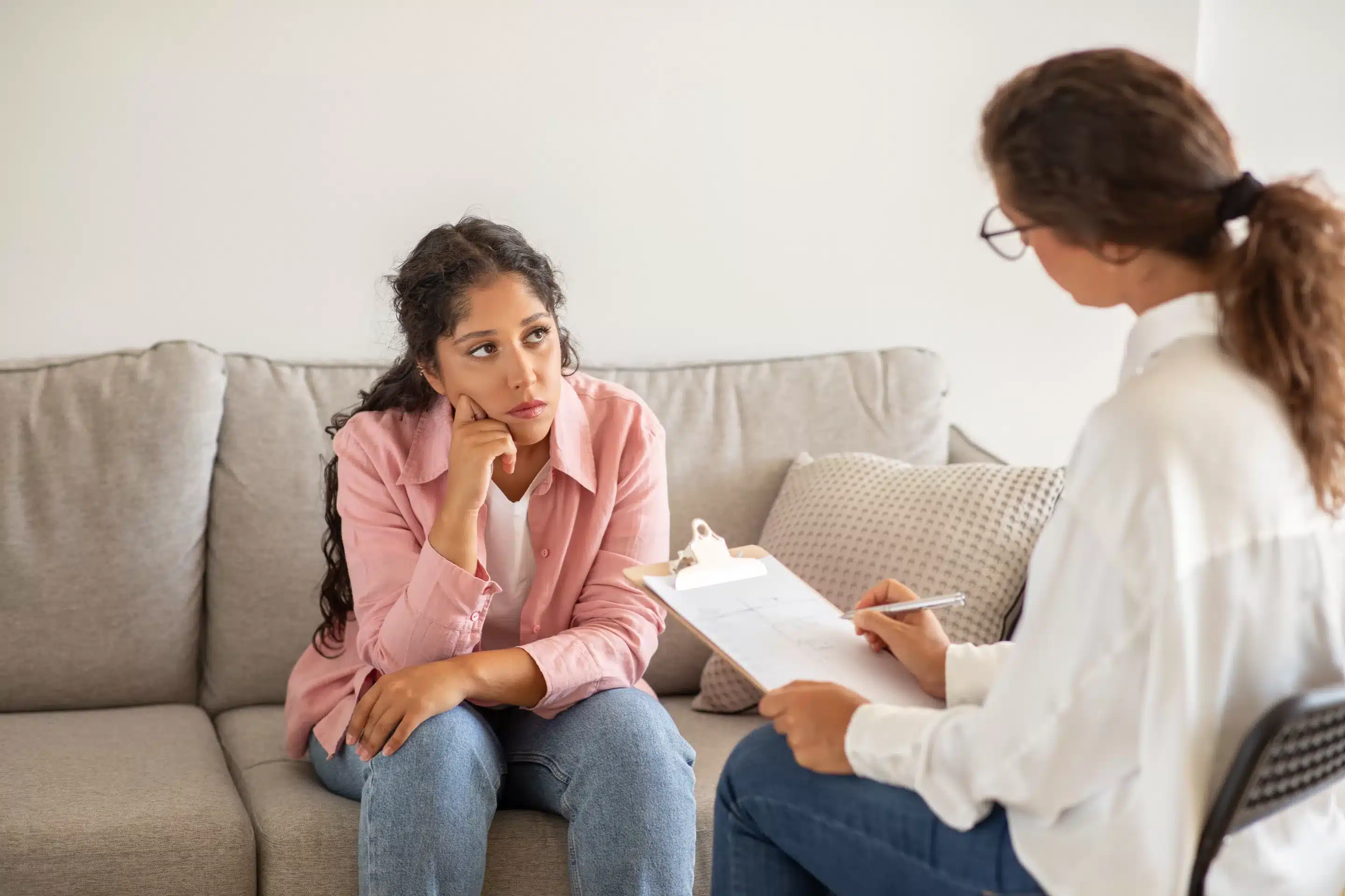 A woman sits on a gray couch in a white room while a therapist listens and takes notes.