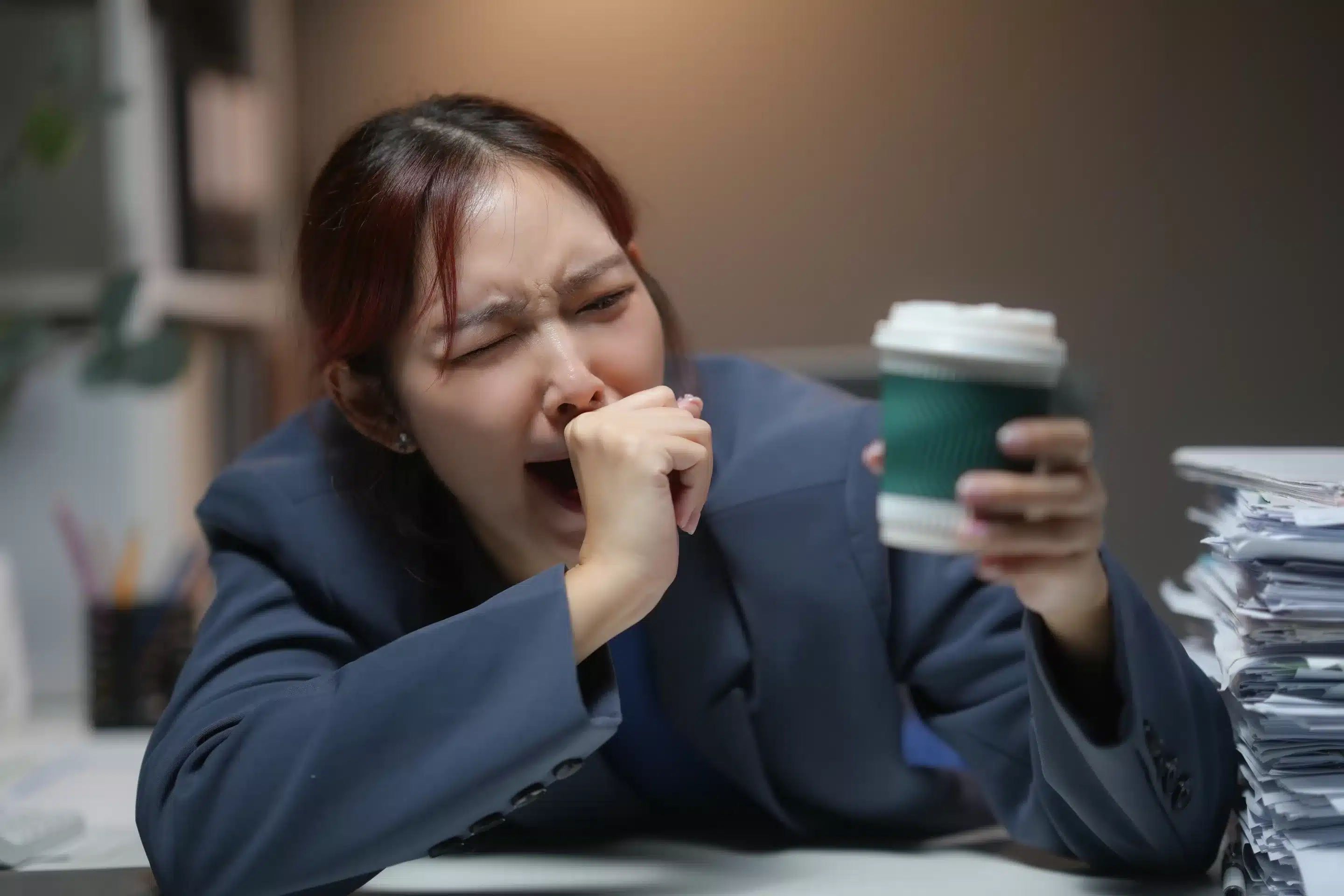 Tired woman in office holding coffee cup, yawning with fatigue,