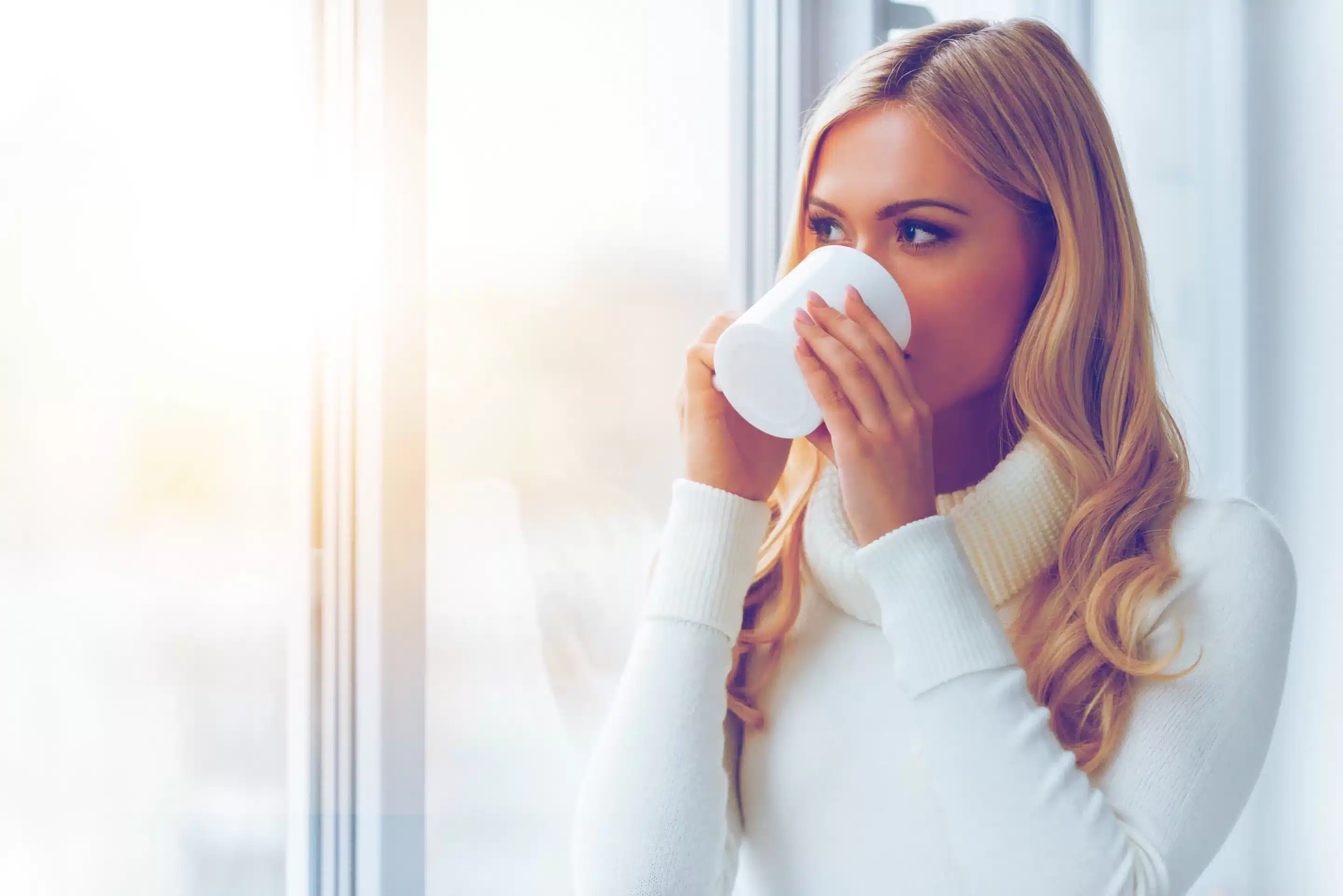 Enjoying fresh coffee. Beautiful young woman in white sweater drinking coffee and looking through a window