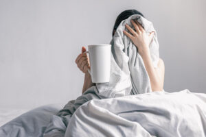 Anxious woman holding coffee cup while lying awake in bed, showing stress