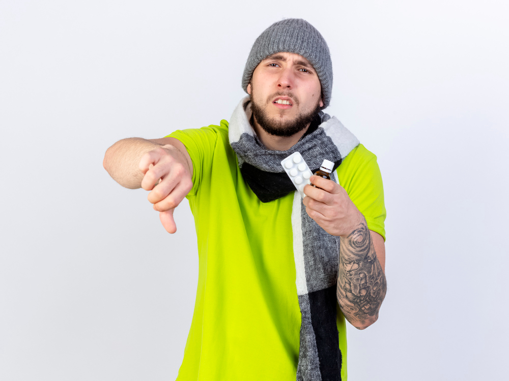 Upset sick young man in winter clothes giving thumbs down while holding medicine