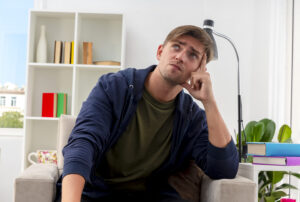 Young blonde man sitting in an armchair with finger on temple, looking up thoughtfully in a cozy living room. How to Focus with ADHD