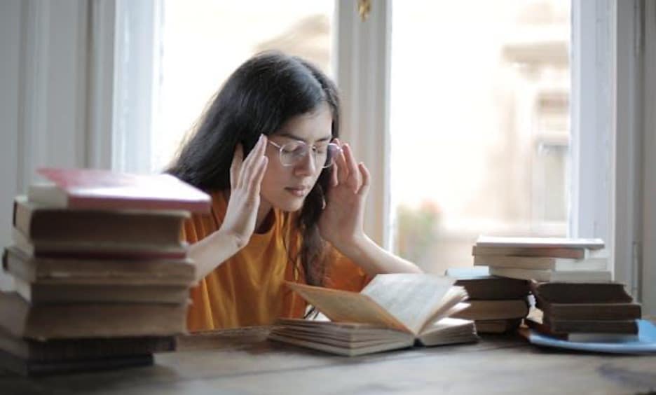 A girl with glasses surrounded by books, trying to focus.